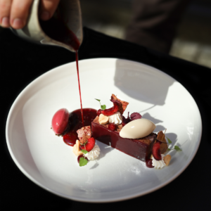 A sophisticated dessert featuring rich berry elements, ice cream, and delicate garnishes, with a red sauce being poured, embodying MICHELIN Guide fine dining presentation